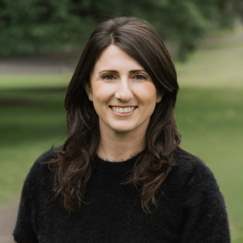 Outdoor headshot of a smiling woman with long dark brown hair, wearing a black top.